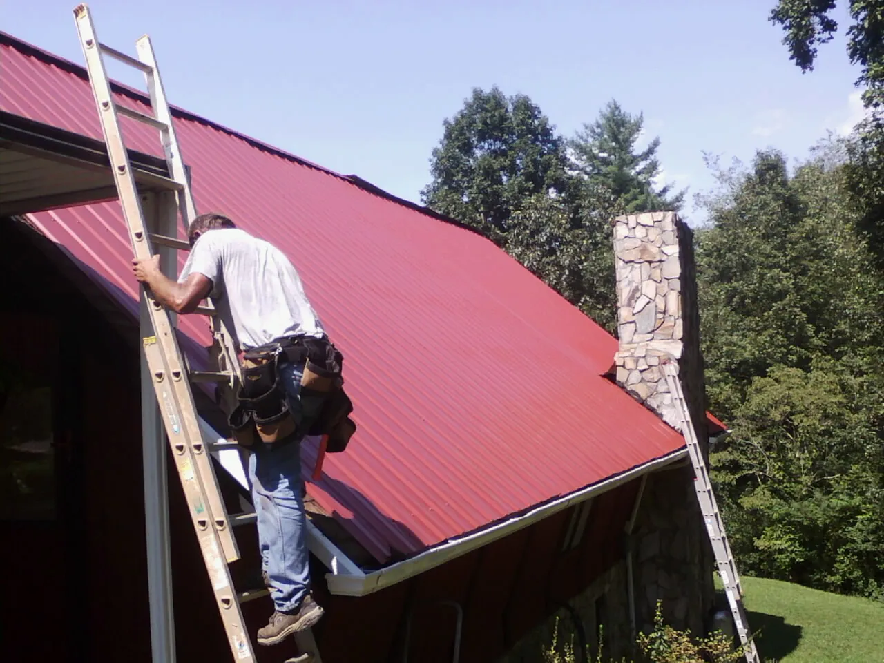 Roofer on a ladder beside a red metal roof and stone chimney — Soffit & Fascia Repair by Roofing Brothers in Schofield